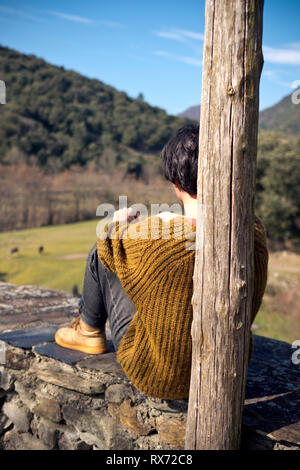 Intime Aufnahme von hinten von Frau mittleren Alters, die auf einer Steinmauer sitzt und über ein Feld mit Eseln und die Berge im Hintergrund blickt Stockfoto