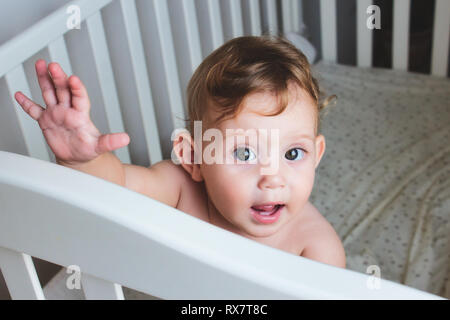 Cute Baby mit Curly blonde Haare stehen in einem Kinderbett, suchen und winken in die Kamera Stockfoto