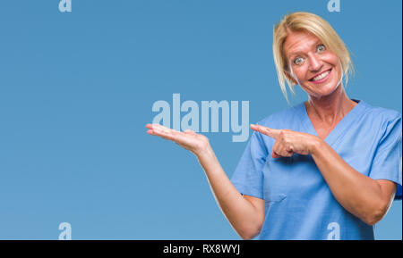 Blonde Frau mittleren Alters tragen Arzt Krankenschwester uniform über isolierte Hintergrund erstaunt und lächelt in die Kamera während der Präsentation und mit der Hand zeigen Stockfoto