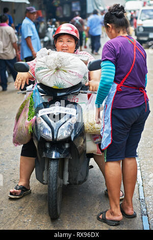 Bangkok, Thailand: Mitte Alter Fett Frau sitzt auf einem Motorrad mit viel Last, die von einem anderen helfen Frau an einem Markt begleitet Stockfoto