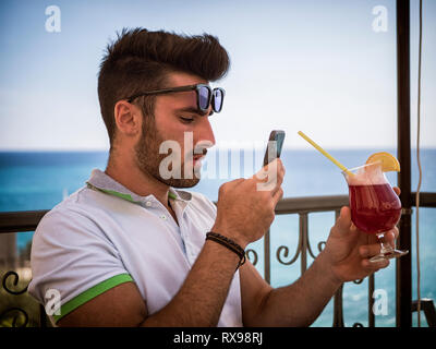 Junge bärtige Mann in Sonnenbrillen chillen auf Resort Terrasse und unter Foto, Cocktail in der Hand Phone Stockfoto