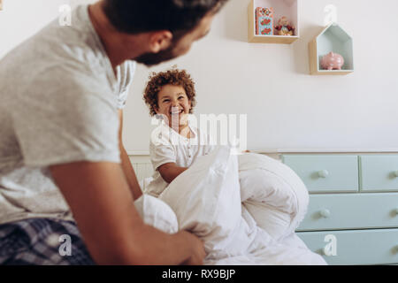 Vater und Sohn in einer Kissenschlacht sitzen auf dem Bett. Fröhlicher Mann und Kind spielt mit Kissen auf dem Bett. Stockfoto