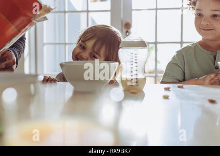 Zwei Kinder am Frühstückstisch sitzen, Milch zu trinken. Man gießt Milch in eine Schüssel geben, während sich die Kinder am Tisch sitzen. Stockfoto