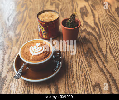 Ein Cappuccino sitzt auf einem Holztisch neben etwas Zucker und ein Kaktus Stockfoto