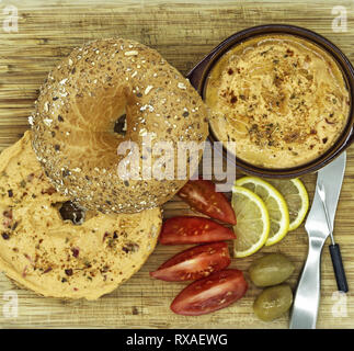 Bagel mit Hummus-Spread auf einem Holzschneidebrett mit einer Dip-Schale, Tomatenscheiben, Oliven und Zitrone, Studio-Food-Shot in einer Flat-Lay-Oberansicht Stockfoto