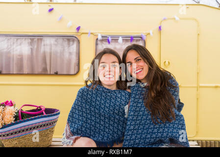 Portrait von lächelnd weibliche Freunde in Decke sitzen gegen gelb Motor home im Hinterhof gewickelt Stockfoto