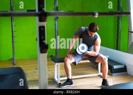 Mann anheben Kurzhantel sitzend Sitz gegen grüne Wand im Fitnessraum Stockfoto