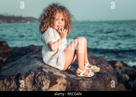 Kleine Mädchen in weißem Kleid auf einem Felsen in der Nähe eines Meeres Stockfoto