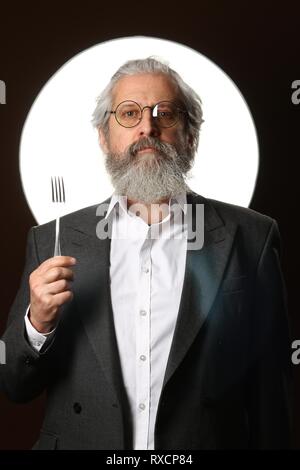 Studio Portrait von ein grauhaariger alter Mann mit einem Struppigen Schnurrbart und Bart, in einem weißen Hemd und graue Jacke, Brille und eine Gabel, auf einem blac Stockfoto