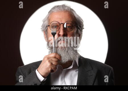 Studio Portrait von ein grauhaariger alter Mann mit einem Struppigen Schnurrbart und Bart, in einem weißen Hemd und graue Jacke, Brille und eine Gabel, auf einem blac Stockfoto