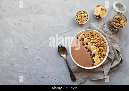 Schokolade smoothie Schüssel mit Bananen, Müsli und Erdnüsse auf Hellgrau konkreten Hintergrund. Gesundes vegetarisches Frühstück. Ansicht von oben mit Platz für Tex Stockfoto