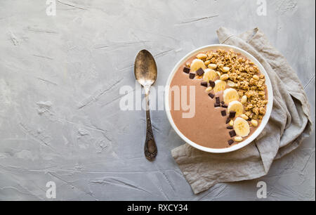 Schokolade smoothie Schüssel mit Bananen, Müsli und Erdnüsse auf Hellgrau konkreten Hintergrund. Gesundes vegetarisches Frühstück. Ansicht von oben mit Platz für Tex Stockfoto