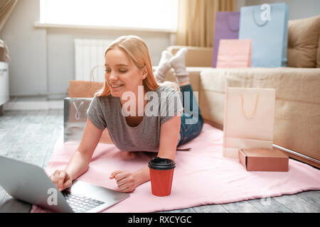 Online Shopping zu Hause. Junge happy Shopper ist auf Laptop und Auswahl von Waren im Online Shop, während auf dem Boden liegend mit Tüten Stockfoto