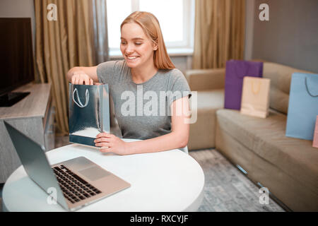 Online Shopping zu Hause. Junge aufgeregt Frau unboxing Ihr Paket, bestellt per Internet, während Sie am Tisch sitzen Stockfoto
