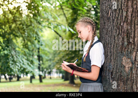 Portrait einer weiblichen Kursteilnehmer Teenager in Uniform mit einem Rucksack, in einem Park in der Nähe eines großen Baumes. Schülerin ein Buch lesen Stockfoto