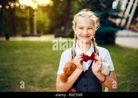 Schülerin mit Zöpfen in Uniformen essen einen Apfel und lächelnd in den Park. Der Student ist eine weiche Bär Spielzeug bei Sonnenuntergang, sie ist Spaß Stockfoto