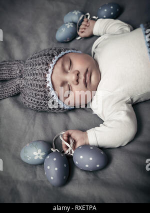 Closeup Portrait von ein süßes neugeborenes Baby tragen Grau gestrickt Kaninchen hat und das Schlafen auf die graue Decke Hintergrund mit Grau festliche Eier, stilvolle E Stockfoto