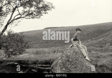 Junge Junge, ca. 7/8 Jahre alt und sitzt auf einem Felsen am West Dart River, Dartmoor, Devon, Großbritannien. Schwarz-weiß Film Fotografie, circa 1994. MODEL RELEASED Stockfoto