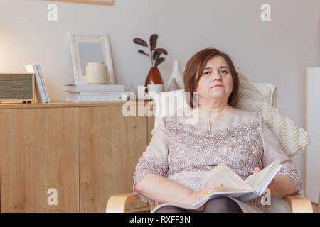 Schöne ältere Frau zurück sitzen auf Sessel mit interessanten Buch. Stockfoto
