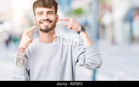 Jungen gutaussehenden Mann mit Sweatshirt über isolierte Hintergrund lächelt zuversichtlich zeigen und zeigen mit den Fingern Zähne und Mund. Gesundheit Konzept. Stockfoto