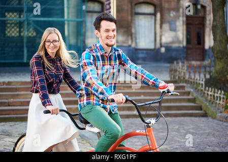 Nahaufnahme der jungen aktiven Lächeln reisenden Paar, gut aussehenden bärtigen Mann und hübsche blonde Frau in Gläsern auf Tandem Bike auf hellen, sonnigen Tag auf St Stockfoto