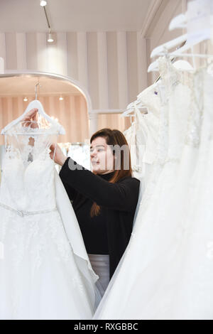 2 Verkäuferinnen helfen, eine hübsche rothaarige Mädchen in einer Wedding Dress Shop Ihr Traum Kleid abholen Stockfoto