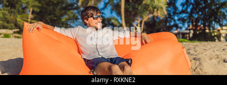Sommer lifestyle Portrait der Mann sitzt auf dem Orange aufblasbare Sofa auf den Strand von tropischen Insel. Entspannen und genießen das Leben auf der Luft Bett BANNER Stockfoto