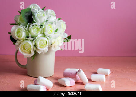 Copyspace mit bunten mini Marshmallows auf einem Tisch neben weißen Rosen auf einem rosa Hintergrund Stockfoto