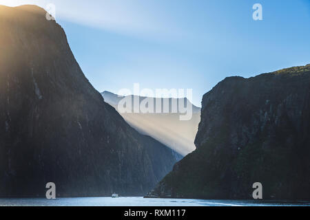 Single Schiff im Milford Sound fjord während der Morgen in Neuseeland. Sonnenstrahlen leuchten über der Oberseite der umliegenden Berge. Stockfoto