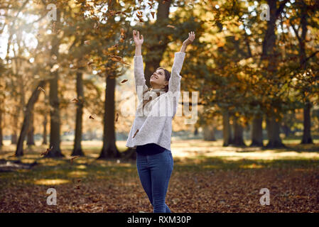 Freudige attraktive junge Frau werfen Herbst in der Luft über dem Kopf Blätter mit einem glücklichen Lächeln im Freien in einem Park mit bunten Herbstfarben auf t Stockfoto