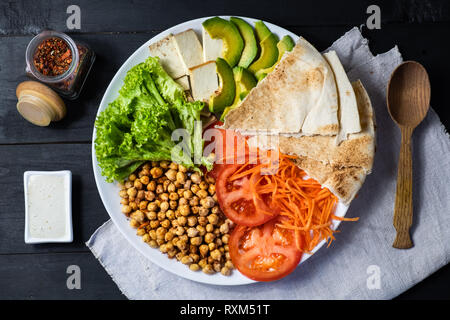 Blick von oben auf die Buddha Schüssel auf einem rustikalen Tisch. Vegane Mahlzeit aus Kichererbsen, Salat, Gemüse, Tofu, Fladenbrot und Avocado, flach Stockfoto