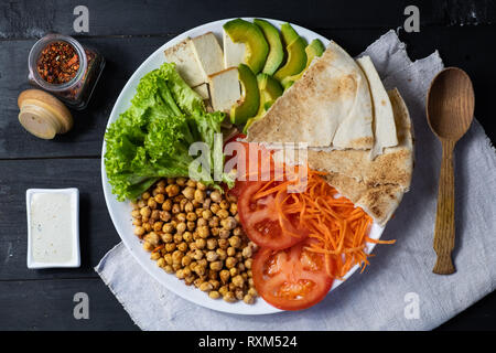 Blick von oben auf die Buddha Schüssel auf einem rustikalen Tisch. Vegane Mahlzeit aus Kichererbsen, Salat, Gemüse, Tofu, Fladenbrot und Avocado, flach Stockfoto