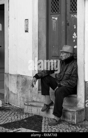 Ein Mann sitzt auf einer Tür, an der Ecke der Rua Capelão und Rua da Mouraria, Martim Moniz, Lissabon, Portugal. Schwarz und Weiss Stockfoto