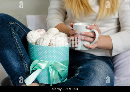 Weibliche Hände halten eine weiße Tasse mit latte Kaffee. Marshmallows amd Baisers in einer Box. Life style Konzept. Nahaufnahme Stockfoto