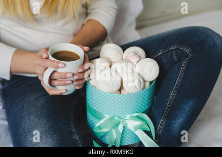 Weibliche Hände halten eine weiße Tasse mit latte Kaffee. Marshmallows amd Baisers in einer Box. Life style Konzept. Nahaufnahme Stockfoto