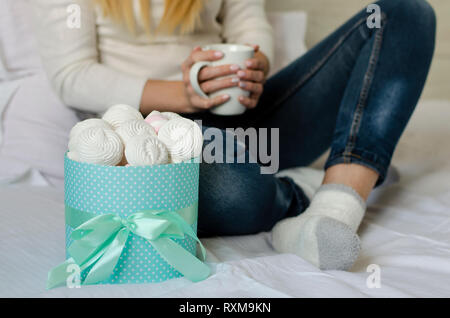 Weibliche Hände halten eine weiße Tasse mit latte Kaffee. Marshmallows amd Baisers in einer Box. Life style Konzept. Nahaufnahme Stockfoto