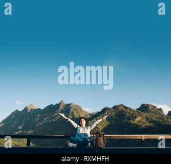 Asiatische Frau Reisende sitzen und die Arme in die Luft an View Point Terrasse an Landschaft, Blick auf die Berge mit Cloud und blauer Himmel im sonnigen Tag an Fores Stockfoto