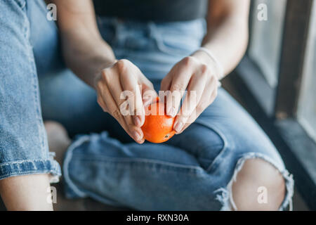 Junge Mädchen Reinigung tangerine sitzen auf der Fensterbank. Nahaufnahme, Hände mit frischen Orange Tangerine. Stockfoto