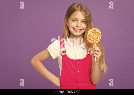 Mädchen mit grossen Candy auf Stick oder Lutscher. Süße Kindheit Konzept. Kind mit langen Haaren gerne Süßigkeiten und Leckereien. Mädchen auf lächelnde Gesicht hält riesigen bunten Lutscher in der Hand, violett unterlegt. Stockfoto