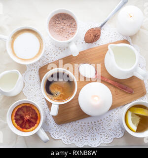 Kaffee, Cappuccino, Kakao und Tee auf einem weißen Tisch mit einem Holzbrett und Kerzen. Ansicht von oben, flach Stockfoto
