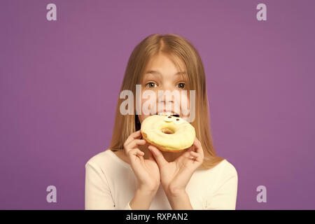 Sweetie Schönheit. Kleines Mädchen essen Donut auf Violett Hintergrund. Kind mit glasierten ring Donut auf lila Hintergrund. Kind mit Junk Food. Essen und Nachtisch. Kindheit und Kinderbetreuung, kopieren. Stockfoto