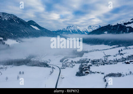 Morgen neblig top Antenne Panoramablick auf Schnee und Dorf mit Bäumen und Straße auf der Winter Landschaft Hintergrund der österreichischen Alpen, Ski Res Stockfoto