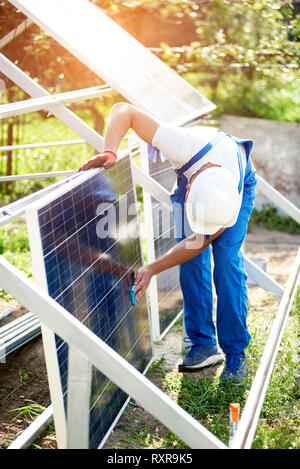 Arbeiter in weißen Helm und Blaumann sauber wischen Big Square glänzende Solarmodule zu Metall Plattform an sonnigen Sommer hell grün Hintergrund lehnte. Stockfoto