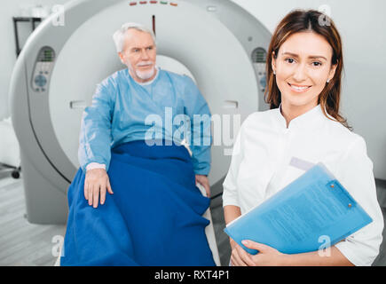 Arzt Radiologe, lächelnd, während bei Kamera schaut. Hinter ihr sitzen ältere Patienten. CT-Scanner im Krankenhaus Stockfoto