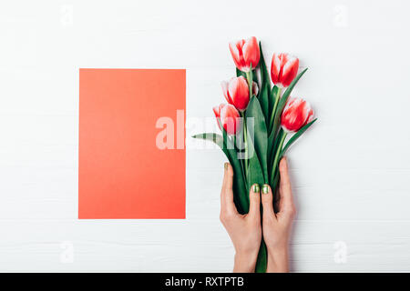 Ansicht von oben Leer rote Karte und Strauß mit frischen Tulpen Blumen in weibliche Hände auf weiße Holztisch, flach. Stockfoto