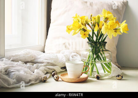 Gemütliches Ostern, Frühling noch leben Szene. Tasse Kaffee, Holzplatte, Wachteleier und Vase mit Blumen auf der Fensterbank. Florale Komposition mit Gelb Stockfoto