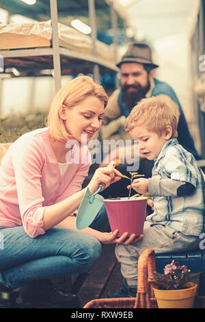 Mutter, die ihren Sohn zarte Blätter der kleinen Blume in Rosa Topf halten Sie Gartenarbeit Spaten. Familie zusammen arbeiten im Gewächshaus Stockfoto