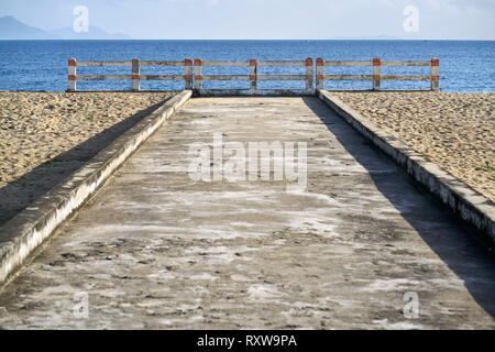 Leere Sandstrand mit einem Gehweg mit einem Zaun auf dem Hintergrund der Meer mit wenigen Boote und Hügel und den Himmel. Sonne scheint. Horizontale. Stockfoto