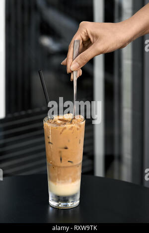 Weibliche Hand einen Löffel im Eis Kaffee trinken mit Eiswürfeln in einem feuchten kalten Glas mit einem schwarzen Stroh auf den dunklen Tisch im Café draußen auf der bl Stockfoto