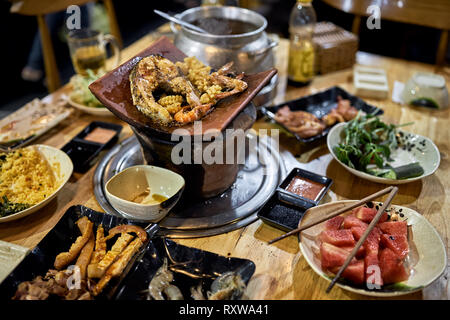 Braten, Fisch, Steak und Krabben auf den heißen Topf mit der Verbrennung von Kohle auf dem Holztisch in der vietnamesischen Cafe. Abwechslungsreiches Essen auf Tellern sind um ihn herum. Clos Stockfoto